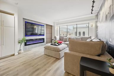 Bright modern urban condo living room with beige sectional and ottoman, ottoman tray with decor, wall-mounted TV with blue LED trim above an electric fireplace, wide city-view windows, light wood floors and potted plant.