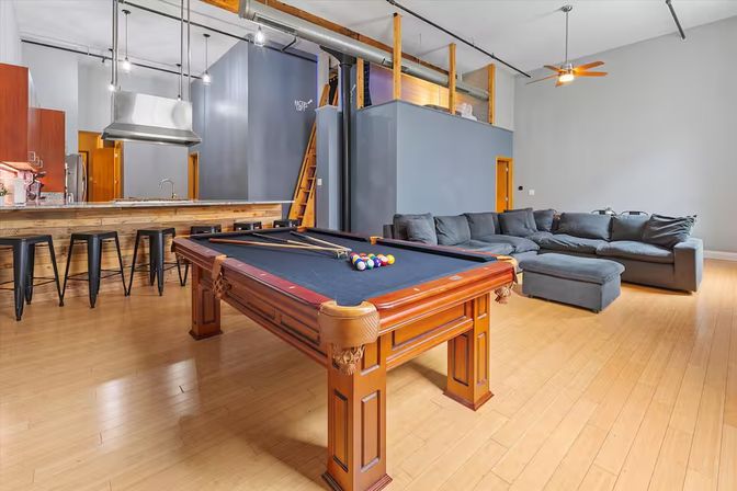 Open-concept urban loft with a wooden pool table (black felt) on hardwood floors, kitchen island with metal barstools and stainless hood, and a large gray sectional sofa beneath high ceilings and exposed ductwork.