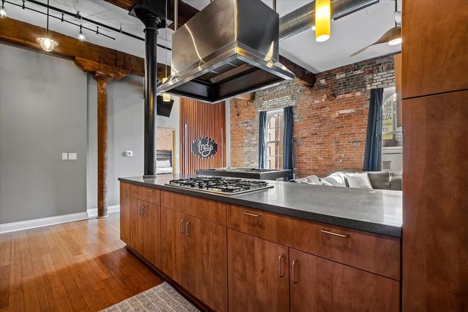 Industrial loft kitchen with exposed red brick and wooden beams, bamboo floors, large gray countertop island with gas cooktop and stainless steel range hood, warm wood cabinets and open-plan living area