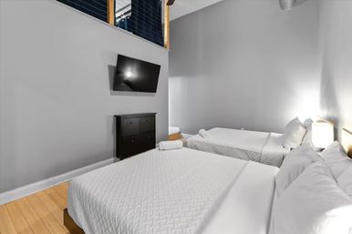 Urban loft-style bedroom with two white quilted beds, gray walls, hardwood floors, wall-mounted flat-screen TV above a dark dresser, bedside lamp and rolled towels.