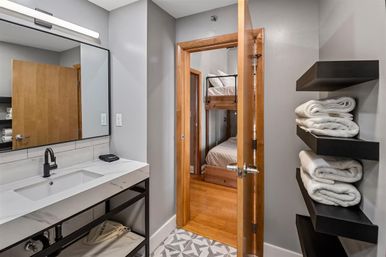Modern gray bathroom with marble-look vanity, black faucet and large mirror, geometric tile floor, black floating shelves stacked with white towels, and an open wooden door revealing a bedroom with wooden bunk beds.