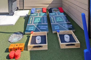 Pair of colorful cornhole drinking-game boards with labeled zones, red and blue bean bags and cup targets on artificial turf backyard patio, ready for a backyard party