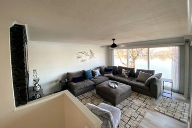 Lakefront living room with large windows, gray L-shaped sectional and ottoman, patterned rug, ceiling fan and tranquil water view.