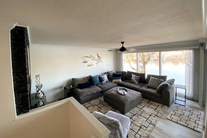 Lakefront living room with large windows, gray L-shaped sectional and ottoman, patterned rug, ceiling fan and tranquil water view.