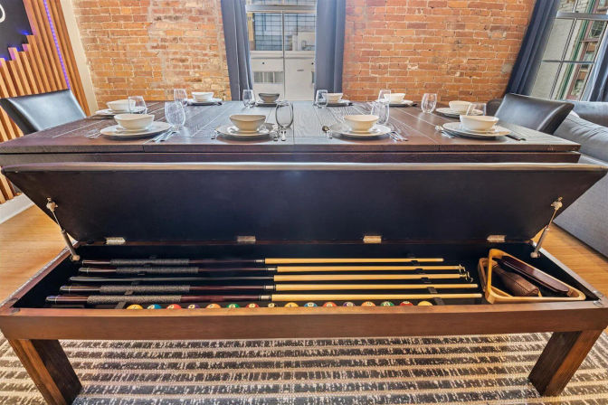Lift-top dining table in an industrial loft with exposed brick and large windows, set for dinner and opened to reveal neatly stored pool cues, billiard balls and triangle rack underneath.