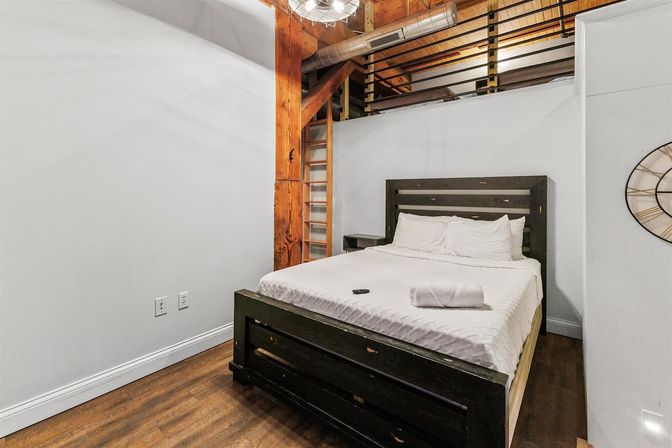 Cozy industrial loft bedroom with a dark wooden bed dressed in white linens and a rolled towel, exposed timber beam, visible metal ductwork, loft railing and warm hardwood floors.