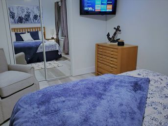Cozy modern bedroom with plush blue throw on the bed, patterned sheets, mirrored sliding closet reflecting a wooden headboard, beige armchair, light-wood dresser with anchor decor and a wall-mounted TV.
