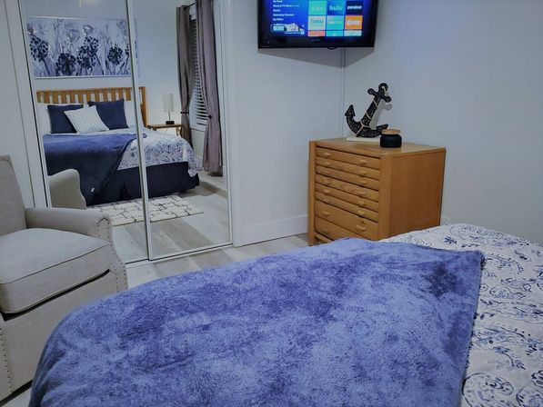 Cozy modern bedroom with plush blue throw on the bed, patterned sheets, mirrored sliding closet reflecting a wooden headboard, beige armchair, light-wood dresser with anchor decor and a wall-mounted TV.