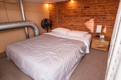 Cozy industrial bedroom with a neatly made bed and white bedding against an exposed red brick wall, metal HVAC ductwork, a wooden nightstand with lamp and alarm clock, and a standing fan.