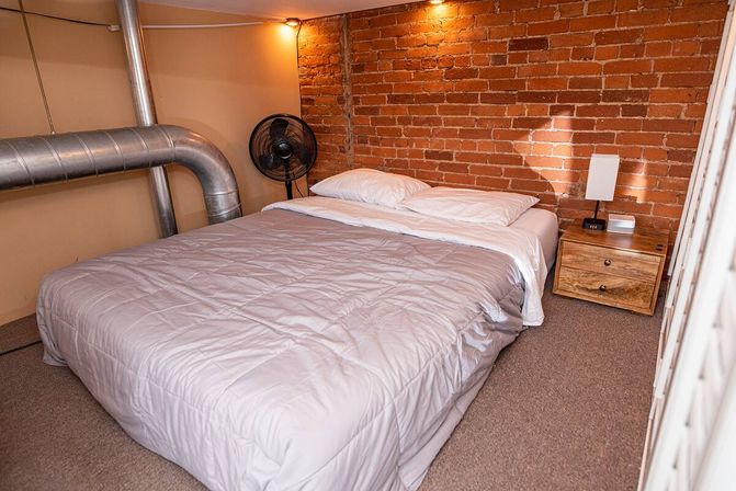 Cozy industrial bedroom with a neatly made bed and white bedding against an exposed red brick wall, metal HVAC ductwork, a wooden nightstand with lamp and alarm clock, and a standing fan.