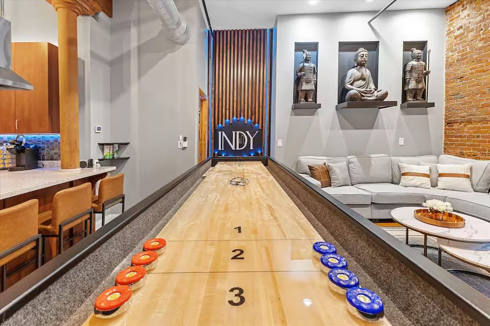 Urban loft game room with a long wooden shuffleboard and red and blue pucks, leather bar stools at a kitchen counter, gray sectional sofa, exposed brick wall and decorative Buddha sculptures.