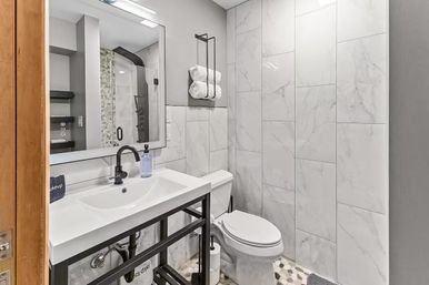 Bright modern bathroom with large white marble wall tiles, black metal-framed vanity and faucet, glass walk-in shower, white toilet, rolled towels on a wall rack, and pebble-pattern floor