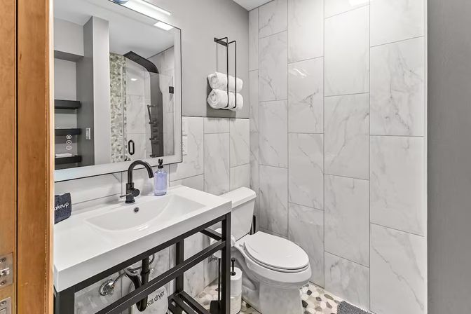 Bright modern bathroom with large white marble wall tiles, black metal-framed vanity and faucet, glass walk-in shower, white toilet, rolled towels on a wall rack, and pebble-pattern floor