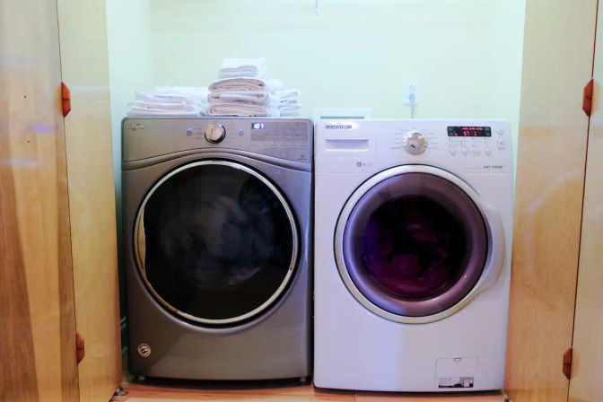 Pair of front-loading washer and dryer in a compact laundry closet—silver dryer and white washer side by side with neatly folded white towels on top, wooden doors and light-colored wall