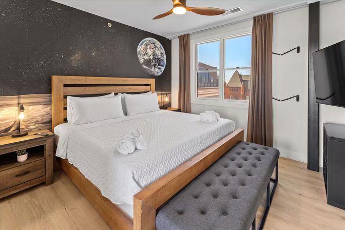 Bright modern bedroom with king-size wooden bed dressed in white linens and towel rolls, starry space mural with a planet above the headboard, tufted gray bench, wooden nightstand with lamp, ceiling fan and large window showing an urban city view.