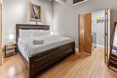 Modern bedroom with king-size bed and white bedding on a dark wood frame, hardwood floors, bedside lamp, framed horned-skull artwork above the headboard, and an open closet with a washing machine and laundry supplies.
