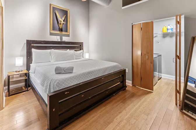 Modern bedroom with king-size bed and white bedding on a dark wood frame, hardwood floors, bedside lamp, framed horned-skull artwork above the headboard, and an open closet with a washing machine and laundry supplies.