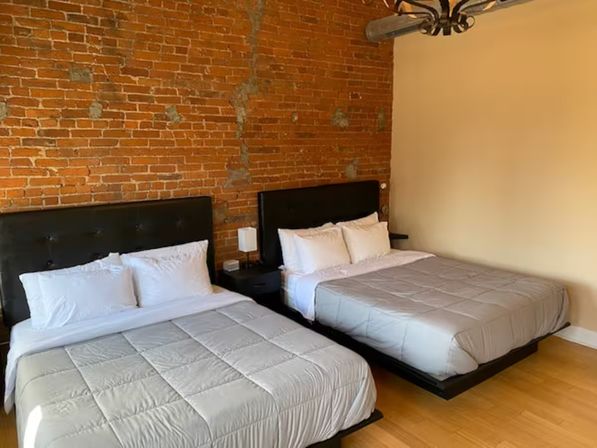 Cozy urban loft-style guest room with exposed red brick wall, two queen beds with dark tufted headboards, white linens and light gray comforters, a central nightstand with lamp, hardwood floors and industrial ceiling fixture