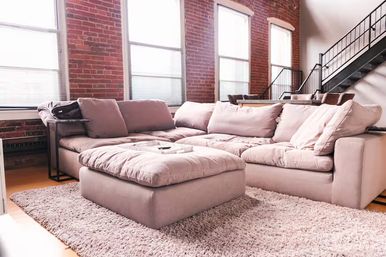 Sunlit modern industrial loft living room with a cozy blush sectional sofa and ottoman on a shag rug, exposed brick walls, tall windows, and a black metal staircase.