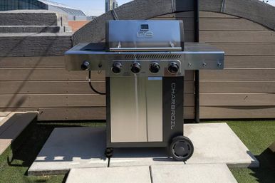 Stainless-steel four-burner gas grill ready for rooftop BBQ on an urban patio with a wood fence, concrete pavers and city buildings in the background.