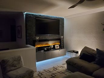 Cozy modern living room with wall-mounted TV above a long electric fireplace showing warm amber flames, cool blue LED backlighting on a low gray media cabinet, plush sectional and accent chair on a patterned rug.