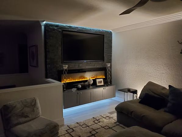 Cozy modern living room with wall-mounted TV above a long electric fireplace showing warm amber flames, cool blue LED backlighting on a low gray media cabinet, plush sectional and accent chair on a patterned rug.
