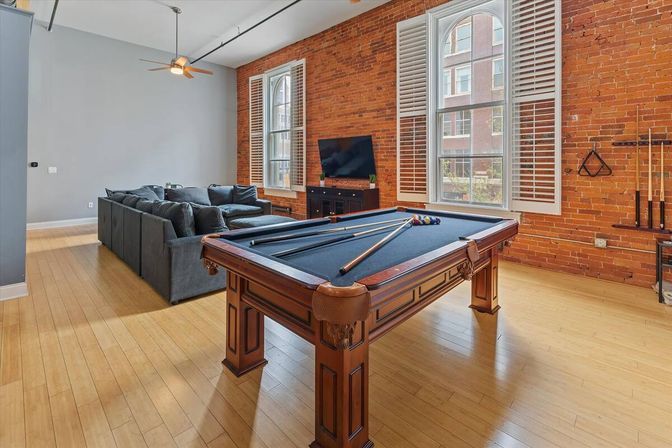 Urban loft game room with a wooden pool table (blue felt) and cues, sectional gray sofa, flat-screen TV, exposed red brick wall with tall arched windows and white shutters, and light hardwood floors.