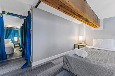 Cozy modern basement bedroom with low ceiling and rustic exposed wooden beam, gray quilted bed with rolled towel, bedside lamp, and blue privacy curtains revealing a second sleeping area.