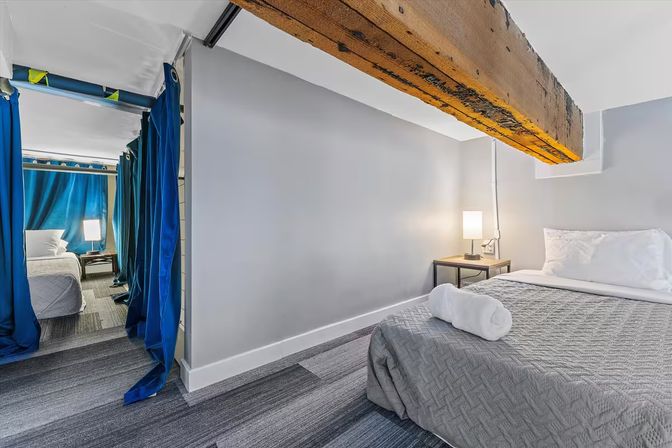 Cozy modern basement bedroom with low ceiling and rustic exposed wooden beam, gray quilted bed with rolled towel, bedside lamp, and blue privacy curtains revealing a second sleeping area.