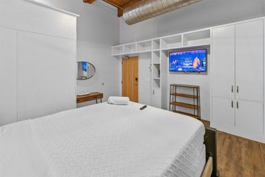 Modern urban loft bedroom with a large white bed, white built-in cabinetry and shelving, wall-mounted TV, wooden ceiling beams and exposed metal ductwork, hardwood floors and a small wooden console.