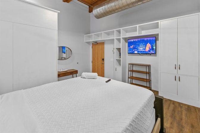 Modern urban loft bedroom with a large white bed, white built-in cabinetry and shelving, wall-mounted TV, wooden ceiling beams and exposed metal ductwork, hardwood floors and a small wooden console.