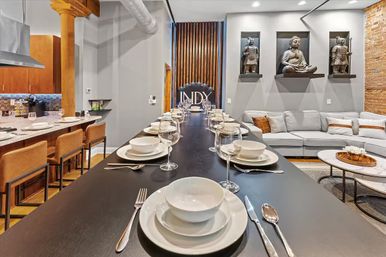 Modern open-concept loft with a long black communal dining table set with white dishes and wine glasses, adjacent kitchen with leather bar stools, exposed wooden column and brick wall, gray sectional sofa, and decorative Buddha and warrior statues on wall shelves.