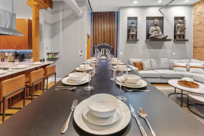 Modern open-concept loft with a long black communal dining table set with white dishes and wine glasses, adjacent kitchen with leather bar stools, exposed wooden column and brick wall, gray sectional sofa, and decorative Buddha and warrior statues on wall shelves.