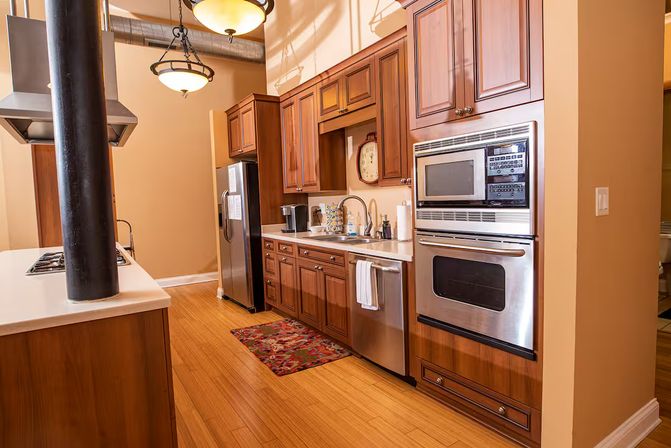 Cozy urban loft kitchen with warm wood cabinets, stainless-steel refrigerator and double oven, island cooktop around a black support column, pendant lights and bamboo floors.