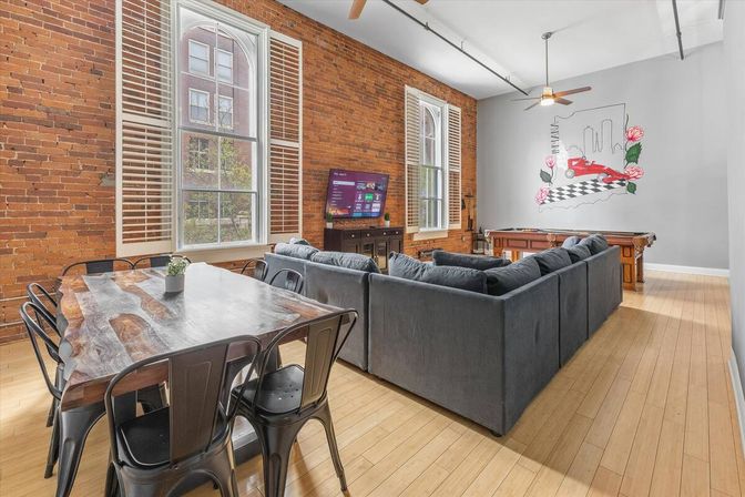 Bright urban loft living area with exposed brick wall, tall arched windows with shutters, hardwood floors, large gray sectional sofa, live-edge wood dining table with metal chairs, wall-mounted TV, pool table and mural.