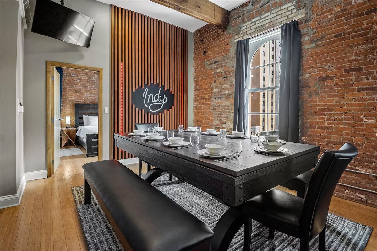Urban industrial loft dining room with exposed brick walls and wooden beam, arched window with gray curtains, black farmhouse table set for eight with white dishes and glassware, bench seating and decorative 'Indy' wall sign.