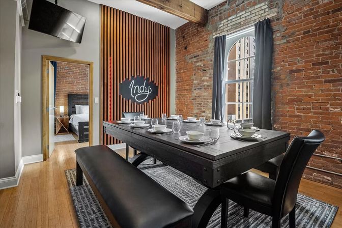 Urban industrial loft dining room with exposed brick walls and wooden beam, arched window with gray curtains, black farmhouse table set for eight with white dishes and glassware, bench seating and decorative 'Indy' wall sign.