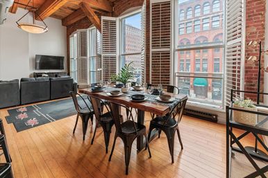 Sunlit industrial urban loft dining area with live-edge wooden table set for six, metal chairs, exposed brick walls and tall plantation-shutter windows overlooking a city street, hardwood floors and open-concept living room with gray sofa.