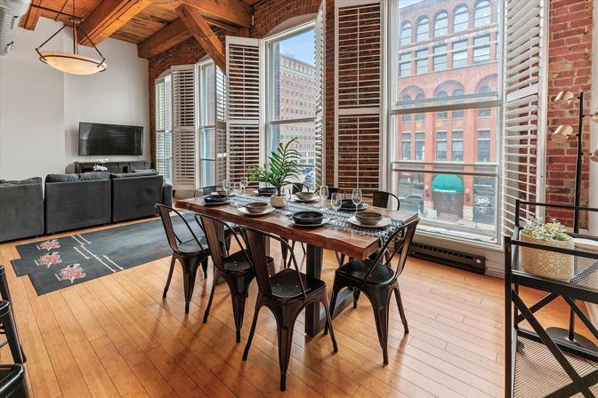 Sunlit industrial urban loft dining area with live-edge wooden table set for six, metal chairs, exposed brick walls and tall plantation-shutter windows overlooking a city street, hardwood floors and open-concept living room with gray sofa.