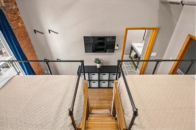Modern urban loft mezzanine bedroom with two beds flanking a central wooden staircase and black metal railings, exposed brick and blue curtain at the window, wall-mounted TV, cube storage and doorway to a tiled bathroom.