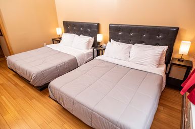 Bright urban hotel room with two large beds, tufted black headboards, white pillows and gray comforters, bedside lamps and hardwood floors