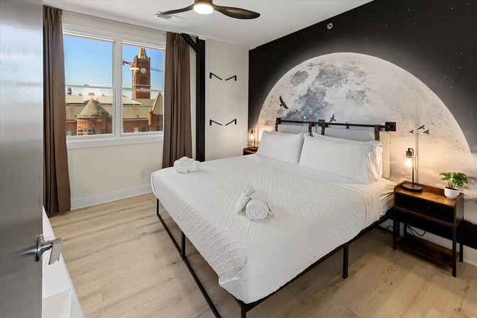 Modern downtown hotel bedroom with a king bed in white linens, oversized moon mural behind the headboard, bedside lamps and a window overlooking a red-brick clock tower.