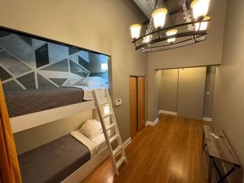 Cozy loft-style bedroom with white built-in bunk beds and ladder, geometric painted accent wall behind the top bunk, warm hardwood floors, industrial chandelier overhead, narrow hallway with small closet and console table.