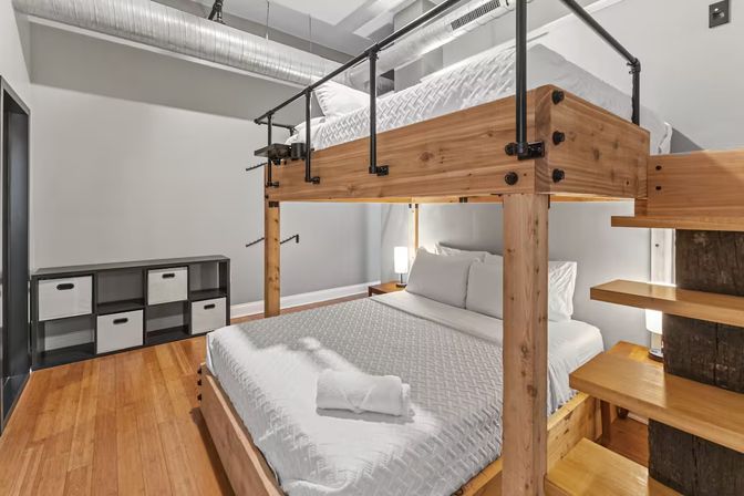 Modern industrial urban loft bedroom with a wooden loft-style bunk bed—lower double bed and upper sleeping platform dressed in white linens, exposed metal ductwork, hardwood floors and cube storage