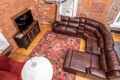 Aerial view of a cozy urban loft living room with exposed red brick walls, hardwood floors, a curved brown leather sectional with cupholders, a vibrant patterned area rug, flat-screen TV on a wooden console, and a white modern floor lamp.