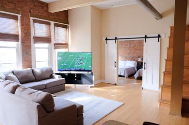 Sunlit urban loft living room with an L-shaped gray sectional, flat-screen TV showing a football game, exposed brick walls, large windows with bamboo shades, white sliding barn doors opening to a bedroom, and light hardwood floors.