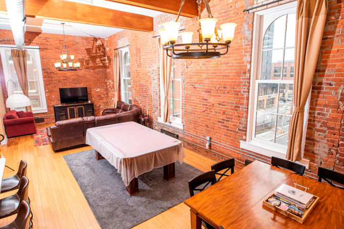Sunlit urban loft with exposed red brick walls and wooden beams, open-plan living and dining area featuring a covered pool table on a gray rug, leather sectional and TV, wooden dining table, chandeliers and large arched windows with a city view.