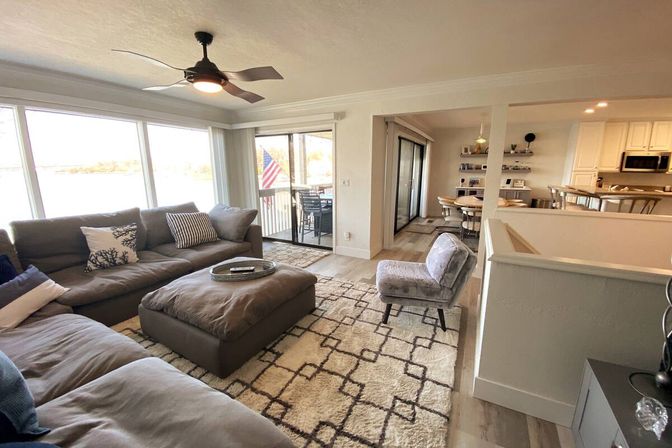 Cozy lakefront open‑concept living room with gray sectional and ottoman, patterned rug, ceiling fan, sliding glass doors to a balcony with an American flag and water views, dining area and kitchen visible