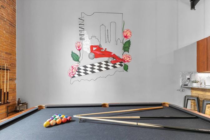 Game room with a black-felt pool table and cues in the foreground and a gray wall mural of Indiana reading "INDIANA" featuring a red Indy-style race car on a checkered flag with floral accents, exposed brick and kitchenette on the sides.
