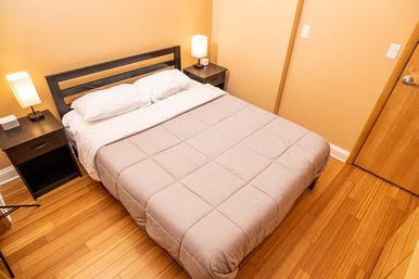 Cozy minimalist bedroom with a double bed in a beige comforter, white pillows, two bedside tables with lamps, warm hardwood floors and tan walls.